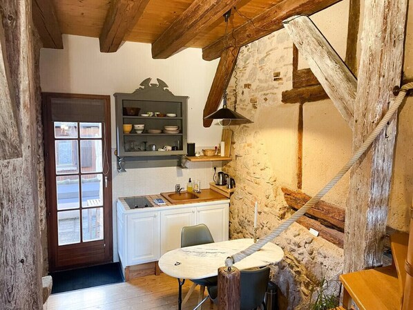 A rustic kitchen with a wooden ceiling, a built-in shelf with dishes, a table with chairs, and a stone wall.