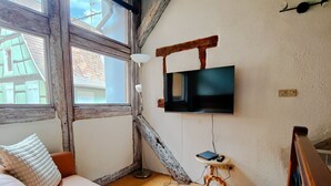 Una habitación acogedora con suelo de madera, un sofá con un cojín blanco, una mesita con una lámpara y un televisor montado en la pared.