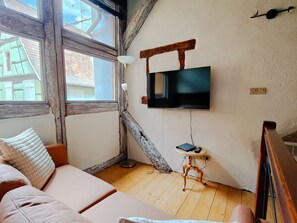 A cozy room with a wooden floor, a sofa with a white cushion, a small table with a lamp, and a TV mounted on the wall.