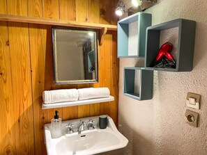 A bathroom with a wooden wall, a white sink, a mirror, and shelves with toiletries.
