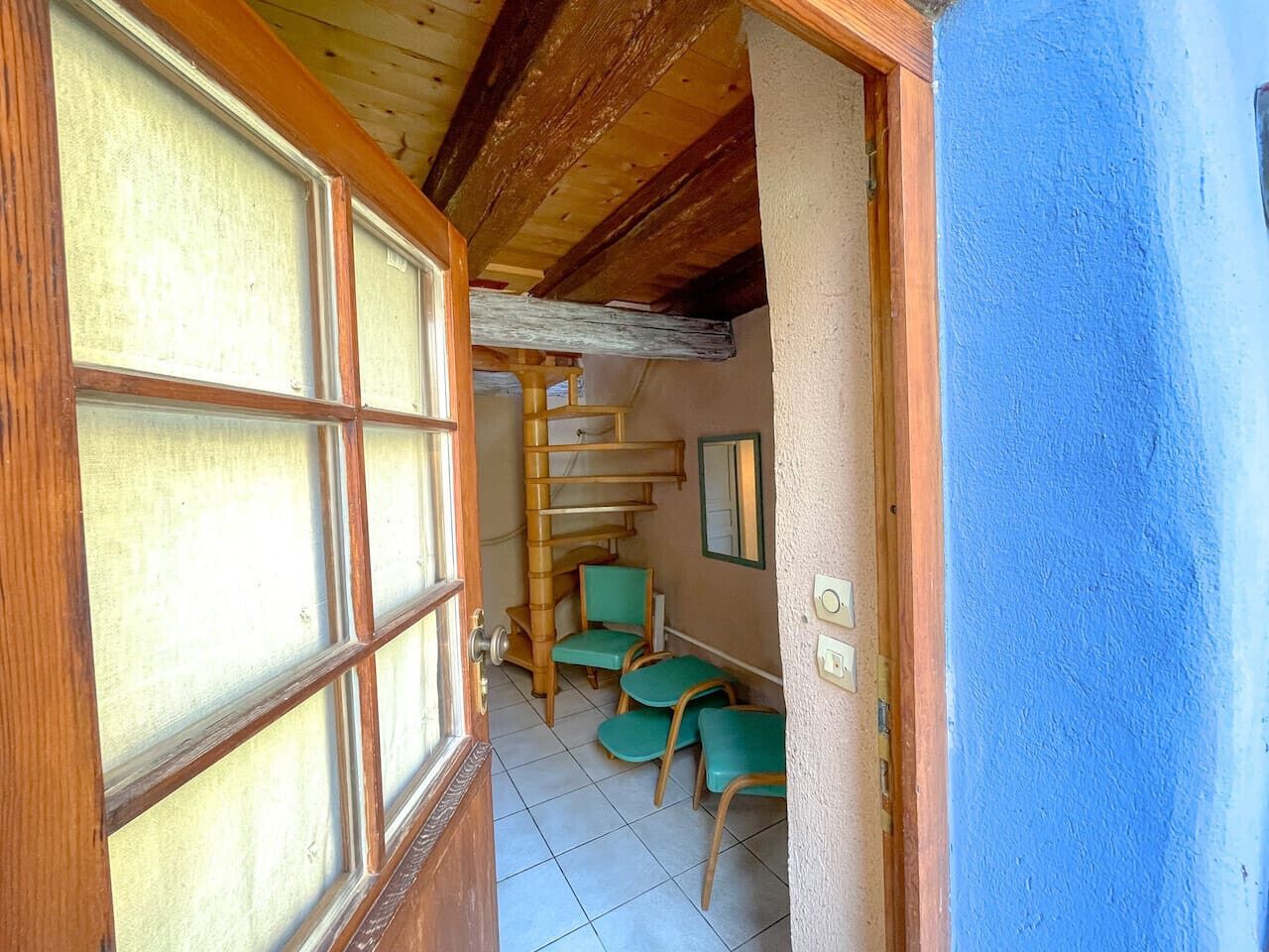 Un couloir étroit avec un escalier en bois, des chaises vertes et un mur bleu.