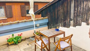 Un conjunto de mesa y sillas de madera dispuesto en un patio con una pared azul y blanca, una cerca de madera y un pequeño jardín con flores.