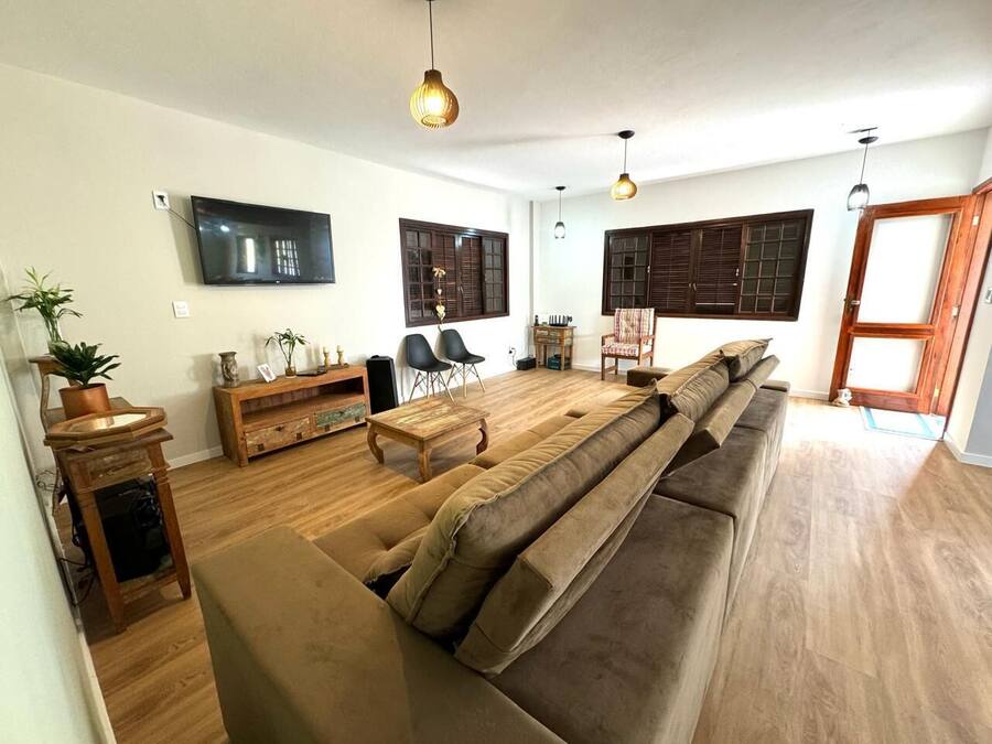 A spacious living room with a large sofa, wooden flooring, a TV mounted on the wall, and a wooden cabinet.