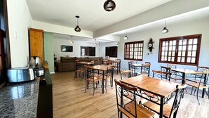 Ruime eetruimte met houten tafels en stoelen, een keuken met een fornuis en koelkast, en grote ramen die natuurlijk licht binnenlaten.