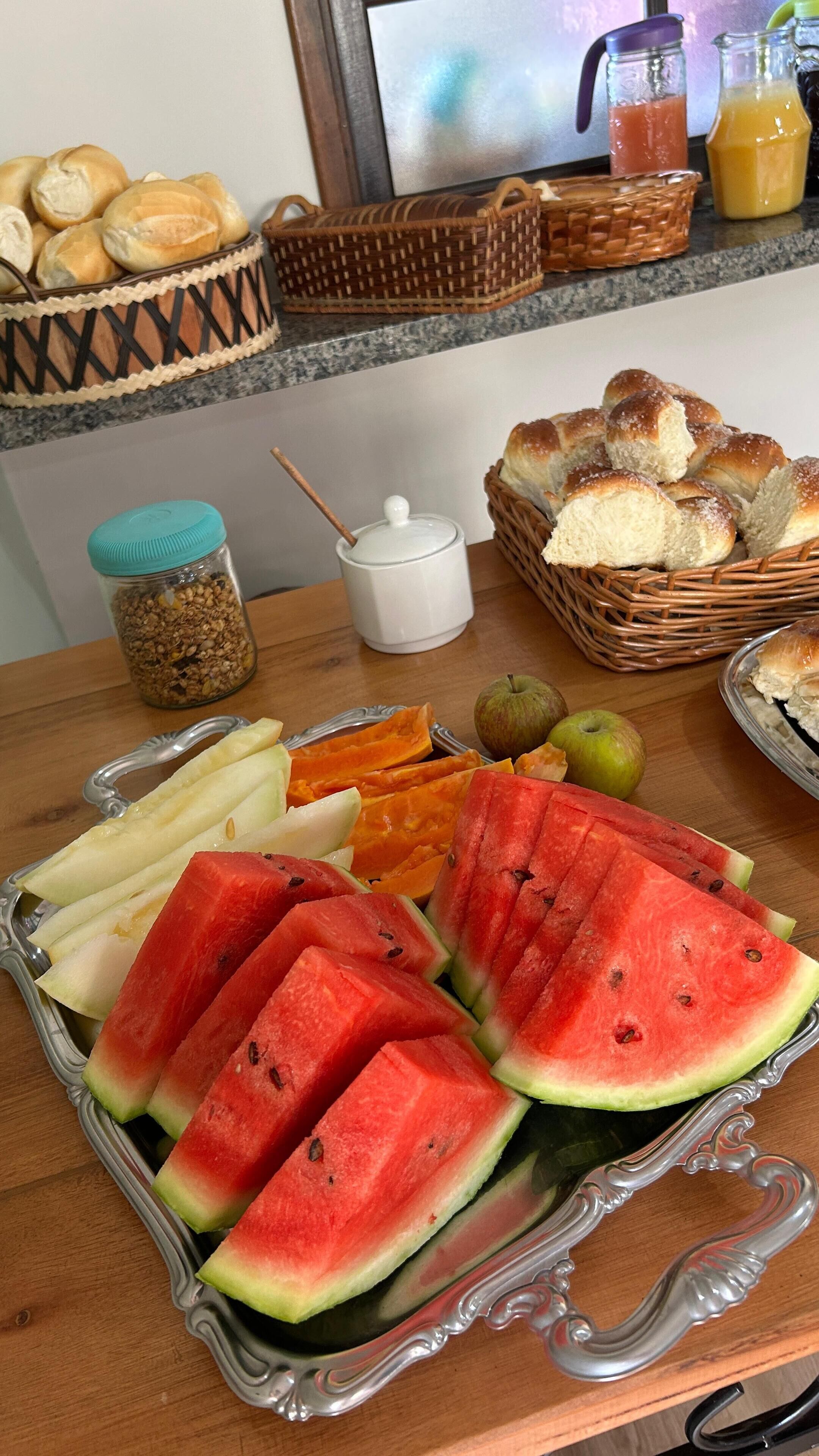 Una mesa de desayuno con sandía, melón y manzanas en rodajas, junto con cestas de pan y un tarro de granola.