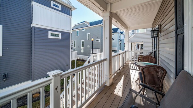 A covered porch with white railings and a wooden deck, featuring outdoor furniture and a view of neighboring houses.