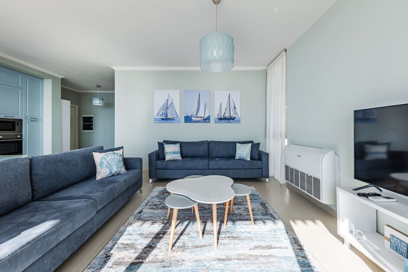 Een moderne woonkamer met een grijze bank, een salontafel en een televisie. Op de muur hangen schilderijen van zeilboten en er is een plafondlamp.