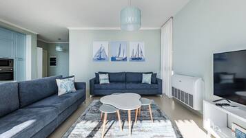 Een moderne woonkamer met een grijze bank, een salontafel en een televisie. Op de muur hangen schilderijen van zeilboten en er is een plafondlamp.