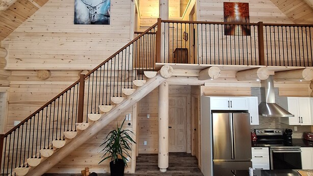 Ruime loft-achtige binnenruimte met een hoge plafond, houten balken, een centrale trap, een keukenhoek en een woonruimte met een bank en een potplant.