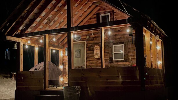 Une cabane en bois avec un porche, illuminée de guirlandes lumineuses, et une pancarte indiquant « Bienvenue ».