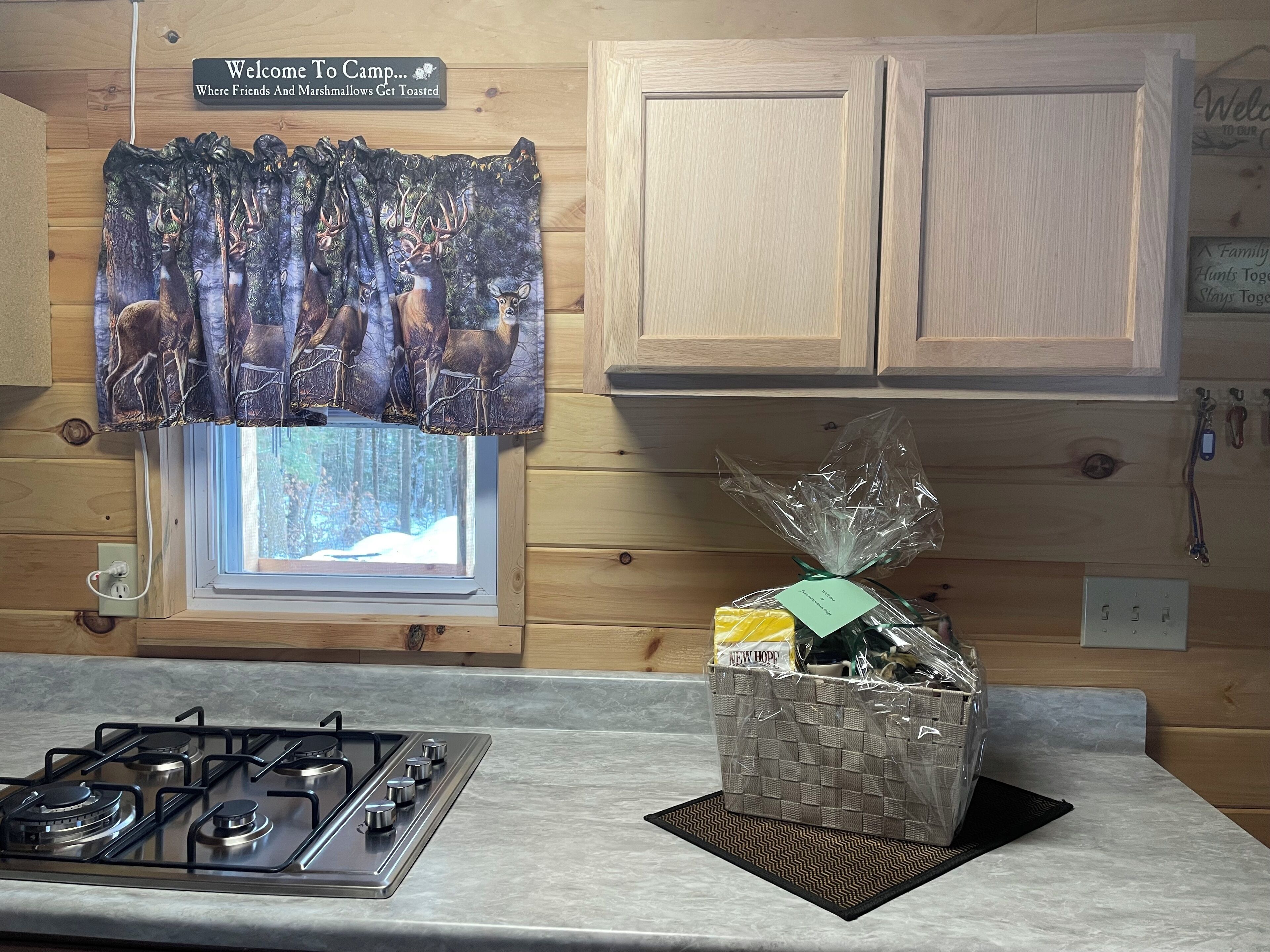 L’intérieur d’une cabane comprend une cuisine équipée d’une cuisinière à gaz, une fenêtre donnant sur une forêt enneigée, des placards en bois et un rideau orné d’un motif de cerf.