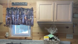L’intérieur d’une cabane comprend une cuisine équipée d’une cuisinière à gaz, une fenêtre donnant sur une forêt enneigée, des placards en bois et un rideau orné d’un motif de cerf.