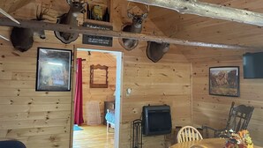 L’intérieur d’une cabane avec des murs en bois, des têtes de cerf accrochées aux murs, une table à manger avec des chaises, un ventilateur de plafond et une cheminée.