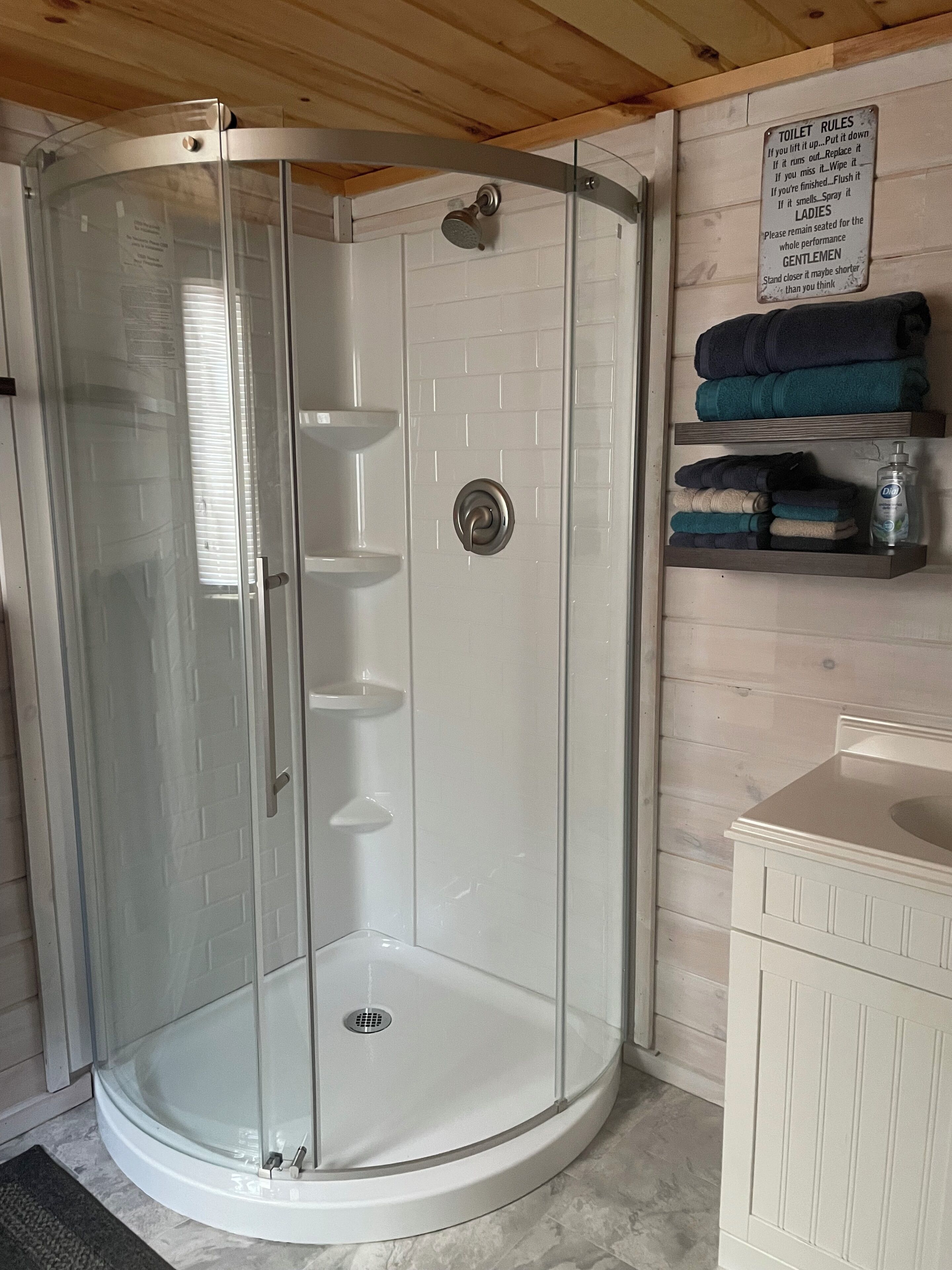 Une cabine de douche ferm ée par des parois de verre, avec un mur recouvert de carreaux blancs, un plafond en bois et un panneau indiquant les règles d’utilisation des toilettes.