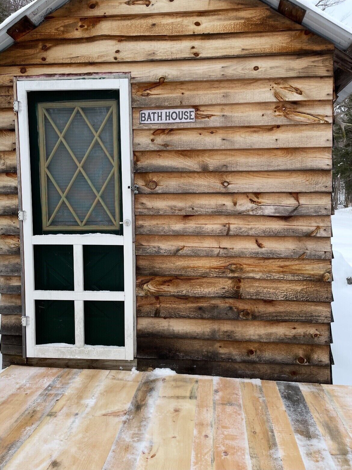 Une cabane en rondins avec une pancarte indiquant « MAISON DE BAINS », une fenêtre à motifs losangés et une terrasse en bois.