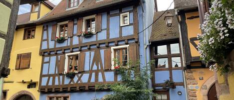 A traditional half-timbered house with a steeply pitched roof, dormer windows, and a small garden area with potted plants and flowers.