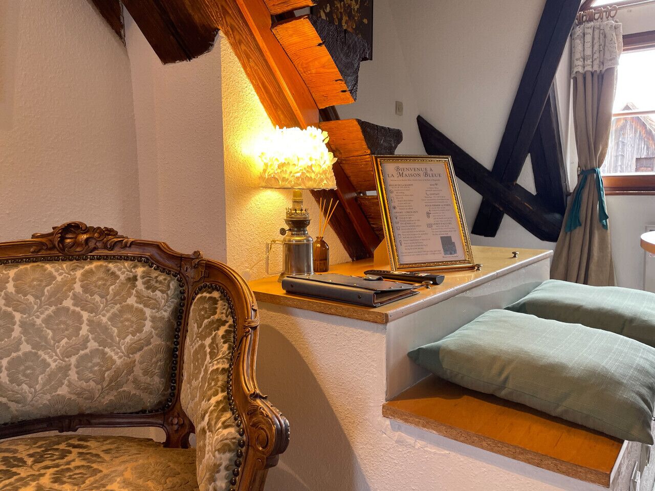 A traditional room with a wooden headboard, a small table with a lamp, a framed welcome sign, and a window with curtains.
