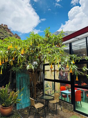 An outdoor seating area with chairs and tables, surrounded by greenery and a mural on the wall. - Anido Hotel (Ollantaytambo)