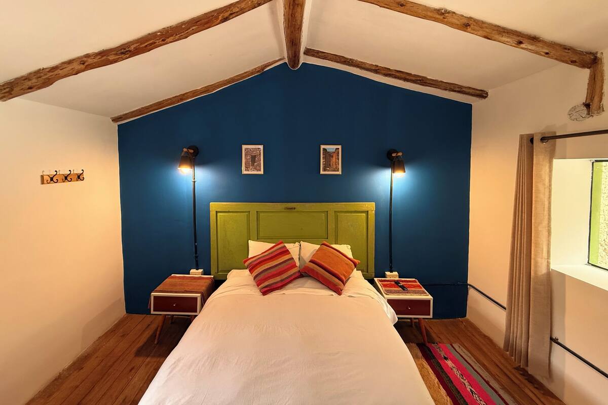 A bedroom with a sloped ceiling, a green headboard, two bedside tables with lamps, a bed with white linens and striped pillows, and a window with curtains.