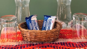A wicker basket containing two chocolate bars, two water bottles, and two glass containers on a table with a red and white patterned tablecloth. - Anido Hotel (Ollantaytambo)