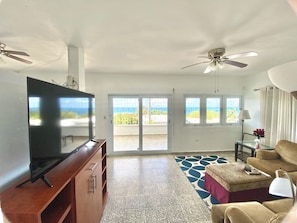 Uma sala de estar com televisão, ventilador de teto, sofá, mesa de centro e vista para o oceano através de amplas janelas.
