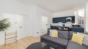 A modern apartment with a grey sofa, a small round table, a kitchen with dark cabinets, a white door, and a potted plant.
