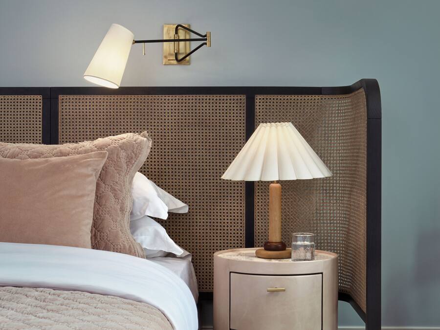 Un letto rifatto con una coperta beige e cuscini, un comodino con una lampada, e un punto luce fissato al muro.
