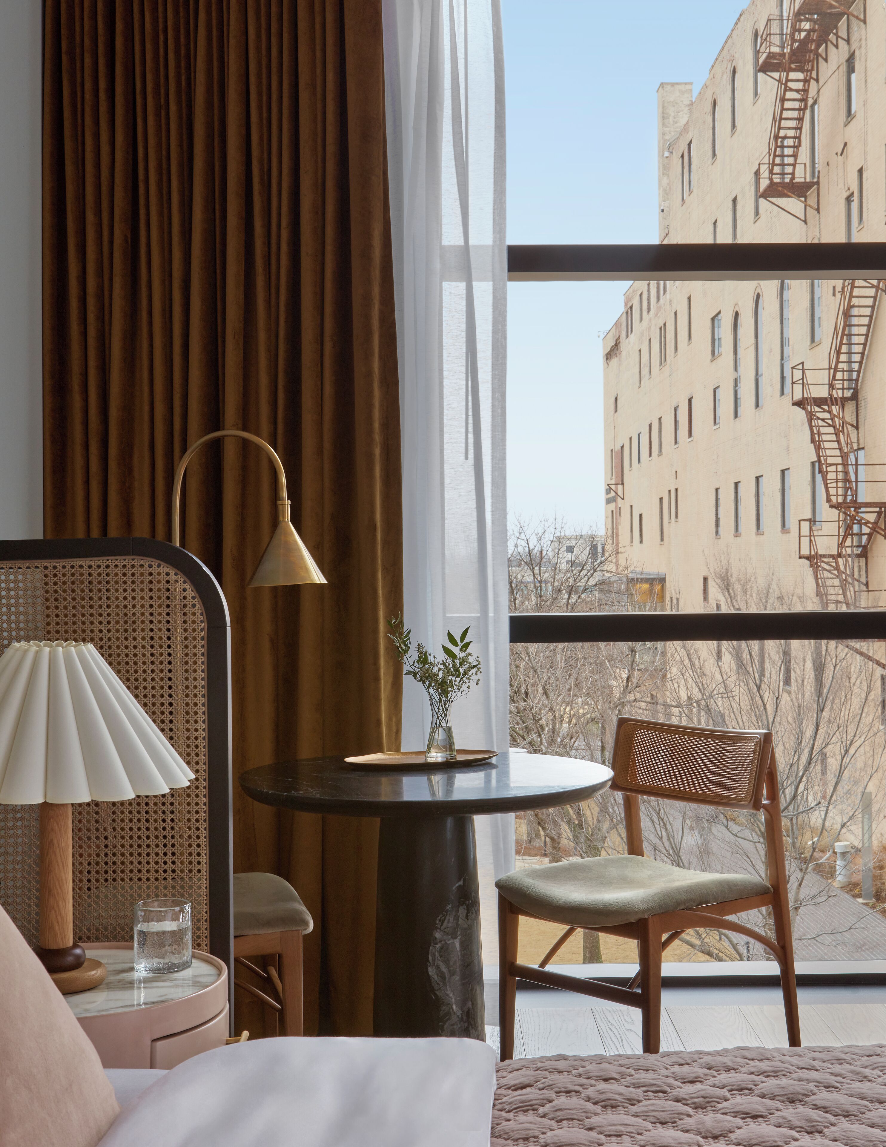 A hotel room with a bed, a round table with a lamp, a chair, and a view of a building through a large window.
