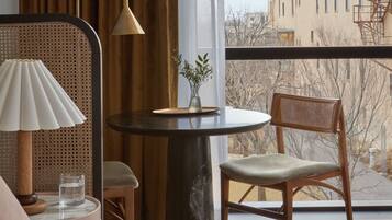 A hotel room with a bed, a round table with a lamp, a chair, and a view of a building through a large window.