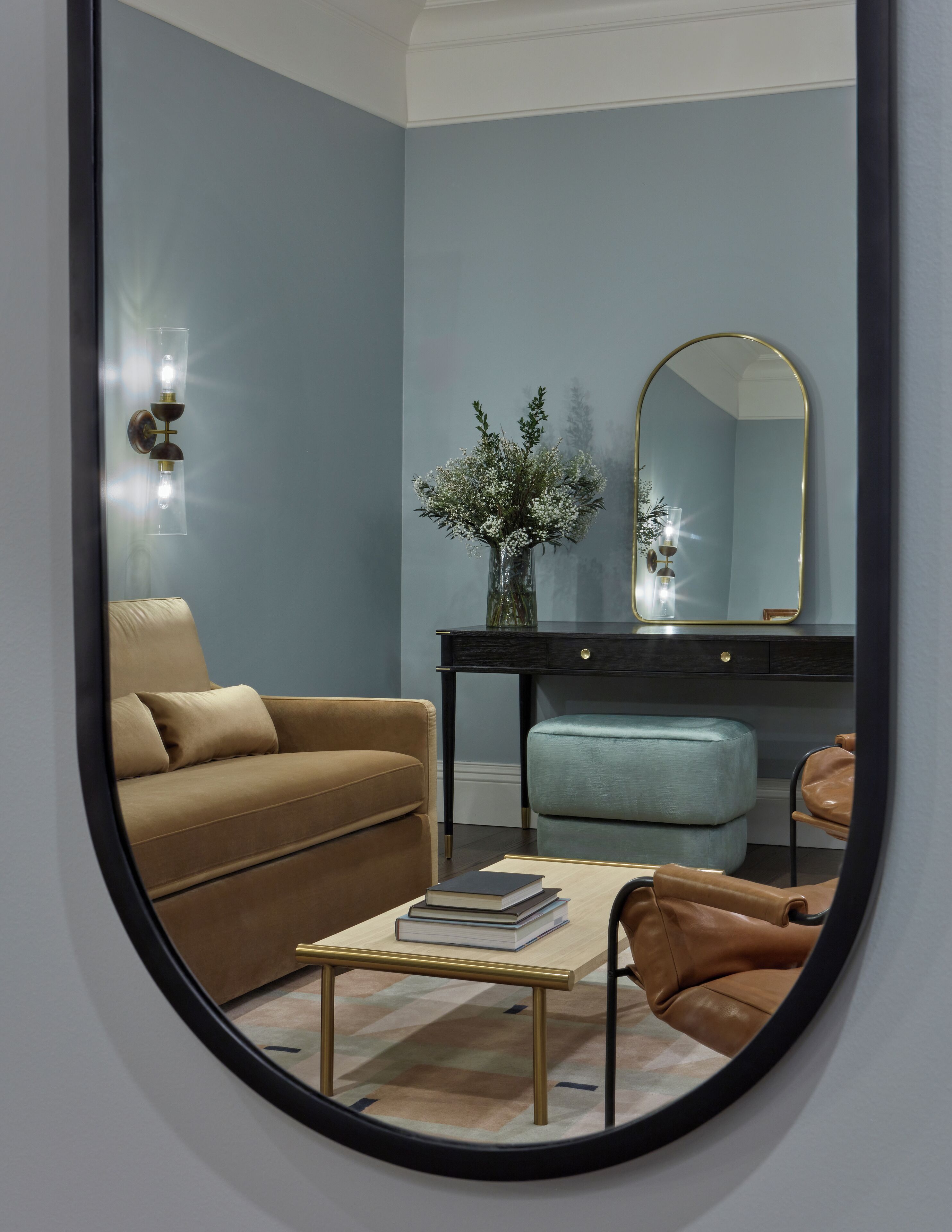 Una habitación con un sofá marrón, un otoman azul claro, una mesita con libros, un espejo y un jarrón con flores blancas.
