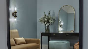 A room with a brown sofa, a light blue ottoman, a small table with books, a mirror, and a vase with white flowers.