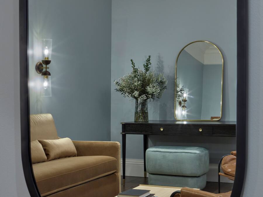 A room with a brown sofa, a light blue ottoman, a small table with books, a mirror, and a vase with white flowers.