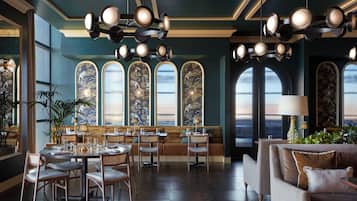 A restaurant with round tables, chairs, and a sofa. There are large windows with decorative panels and a view of the outside.