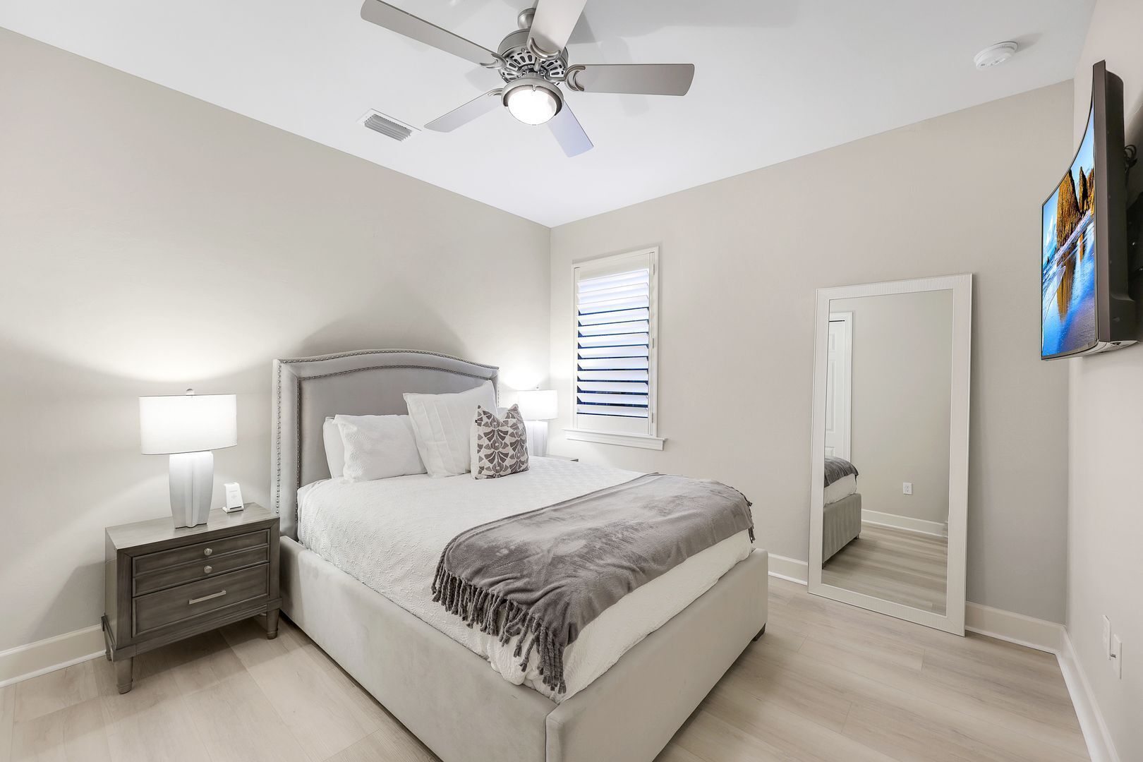 A bedroom with a bed, nightstand, lamp, ceiling fan, window with blinds, and a TV mounted on the wall.