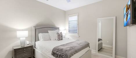 A bedroom with a bed, nightstand, lamp, ceiling fan, window with blinds, and a TV mounted on the wall.