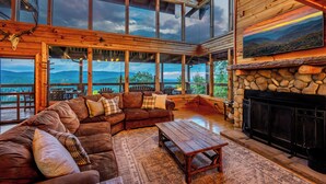 A spacious living room with a large wooden sectional sofa, a stone fireplace, and large windows offering a mountain view. - Serenity - Mountain VIEWS From Every Room! - 71 Windows (Sevierville)