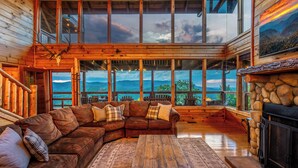 A wooden cabin interior with a large window overlooking a mountainous landscape, a leather sofa, and a stone fireplace. - Serenity - Mountain VIEWS From Every Room! - 71 Windows (Sevierville)