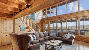 A cozy cabin interior with a wooden staircase, a large window offering a scenic view, and a comfortable seating area with a leather armchair and a wooden coffee table. - Serenity - Mountain VIEWS From Every Room! - 71 Windows (Sevierville)
