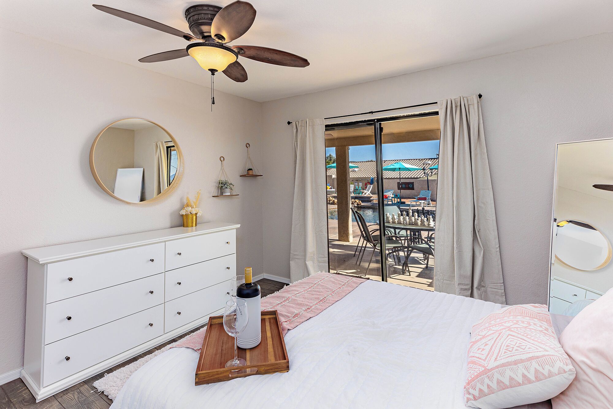 Una camera d'albergo con un letto, una cassettiera, una ventola a soffitto, una finestra con tende e vista su una zona piscina all'aperto.