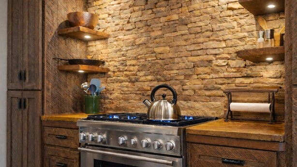 A kitchen with a stone wall, wooden cabinets, a gas stove, and a kettle. - Creek Haüs: Cozy Retreat w/ Endless Entertainment • HotTub • FirePit • GameRoom (Blue Ridge)