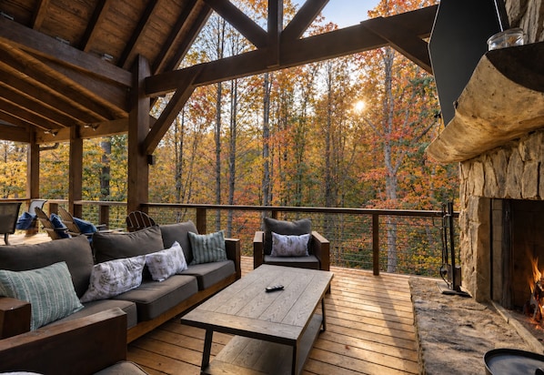 A covered outdoor seating area with a fireplace, wooden deck, and a view of autumn trees.