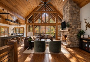 A spacious living area with a stone fireplace, wooden ceiling beams, and a large window overlooking a forest. - Creek Haüs: Cozy Retreat w/ Endless Entertainment • HotTub • FirePit • GameRoom (Blue Ridge)