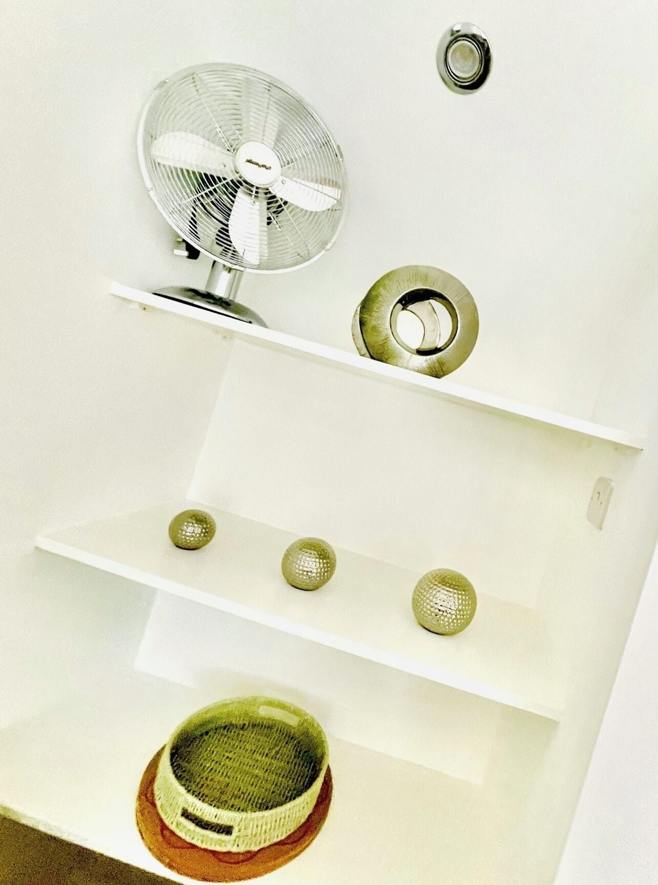 Un mur blanc avec un ventilateur de plafond, un interrupteur et trois étagères ornées d’objets décoratifs.