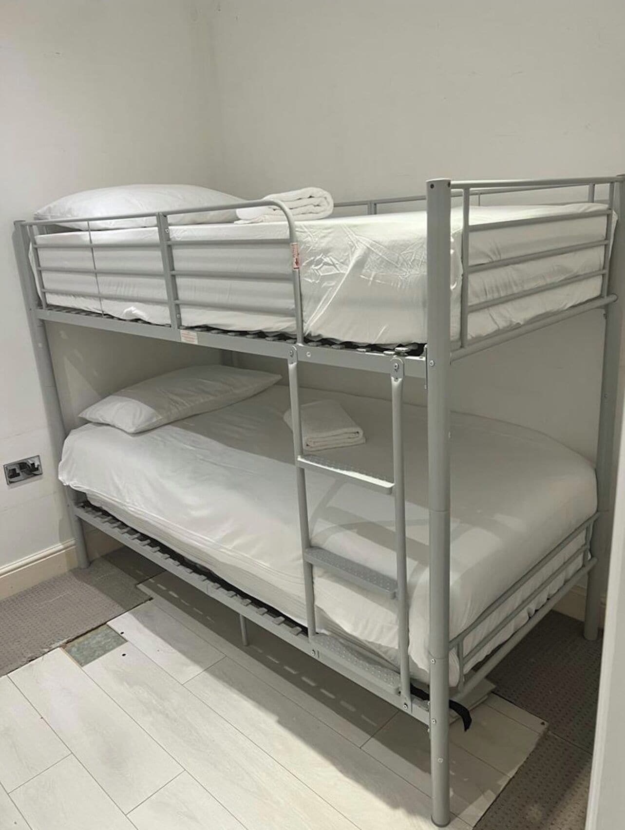 A bunk bed with two levels, each with a mattress and pillows, in a room with light-colored flooring and a plain wall.