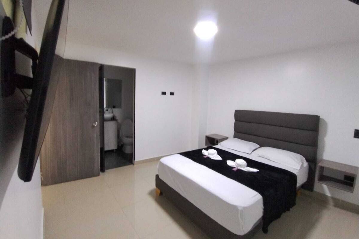 A modern hotel room with a large bed, bedside tables, a wardrobe, and a bathroom visible through an open door.