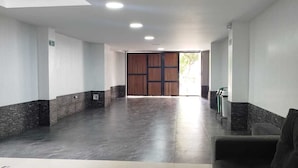 En rymlig entréhall med högt tak, infälld belysning, ett mörkt golv och en stor trädörr längst in.