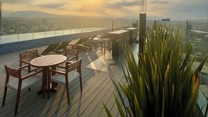 Une terrasse sur le toit, avec une table et des chaises, un pot de fleurs, et une vue sur le paysage urbain et les montagnes au loin.