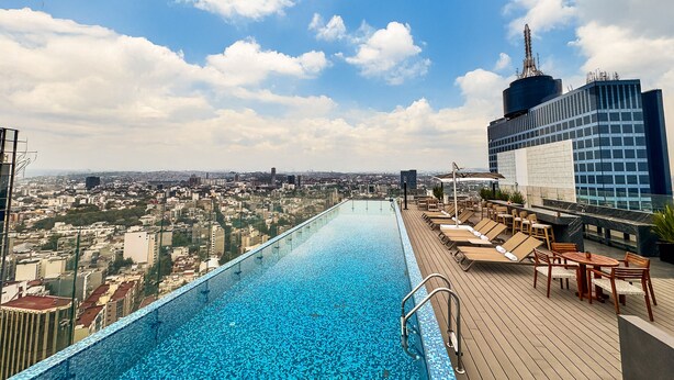 Une piscine à débordement située sur le toit, offrant une vue sur la ville, ainsi que des chaises longues et un espace repas.