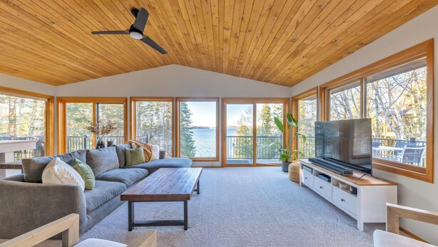 A spacious living room with a wooden ceiling, large windows, a sofa, a coffee table, and a TV on a white cabinet. - Beautiful home on Lake Pokegama! (Grand Rapids)