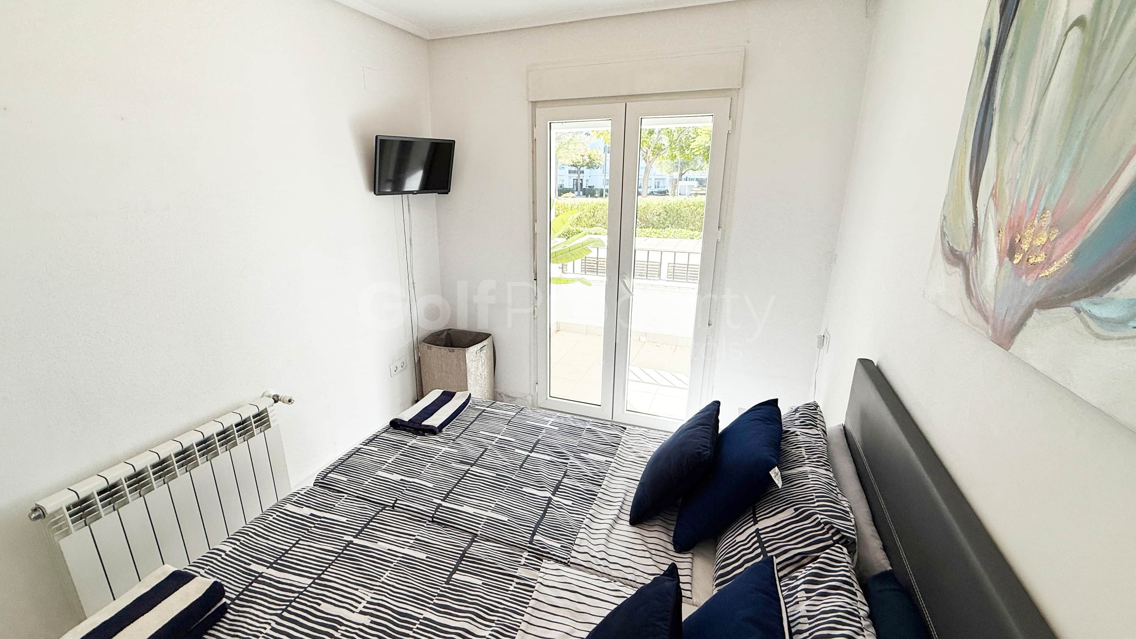 A small, bright room with a bed, a radiator, a TV, and a sliding glass door to an outdoor area.